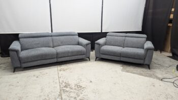 Two grey fabric sofas, a 2-seater and a 3-seater, with contemporary design and slim black legs, placed in a showroom setting.